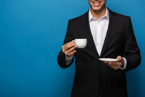 Vista recortada de un hombre de negocios sonriente sosteniendo una taza de café y un platillo sobre un fondo azul - foto de stock