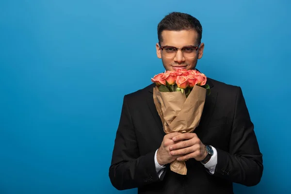 Joven hombre de negocios sosteniendo ramo de rosas y mirando a la cámara sobre fondo azul - foto de stock