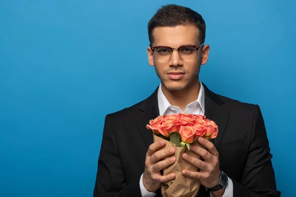 Handsome businessman holding bouquet of roses and looking at camera isolated on blue — Stock Photo