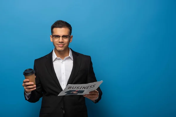 Hombre de negocios guapo sosteniendo periódico y taza desechable sobre fondo azul - foto de stock