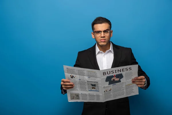 Nachdenklicher Geschäftsmann blickt in die Kamera, während er Zeitung auf blauem Hintergrund hält — Stockfoto