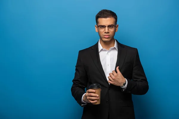 Jovem empresário em óculos olhando para a câmera enquanto segura café para ir no fundo azul — Fotografia de Stock