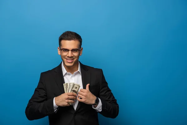 Улыбающийся бизнесмен кладет долларовые купюры в карман пиджака на синем фоне — стоковое фото