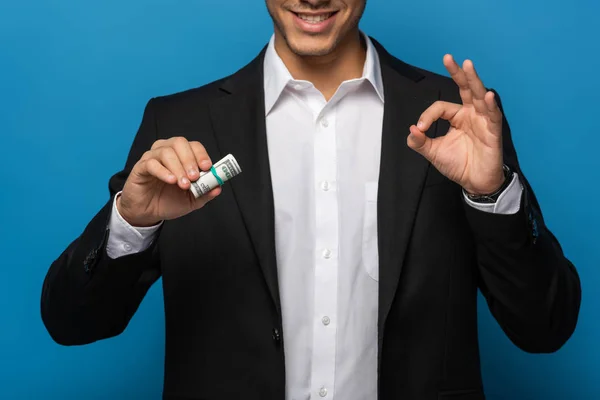 Vista recortada de un hombre de negocios sonriente mostrando un gesto aceptable y sosteniendo el rollo de efectivo sobre fondo azul - foto de stock