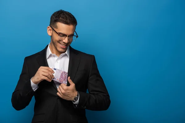 Усміхнений бізнесмен кладе доларові банкноти в кишеню куртки на синьому фоні — стокове фото