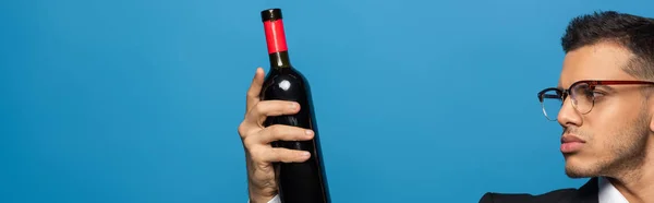Side view of businessman looking at bottle of wine isolated on blue, panoramic shot — Stock Photo