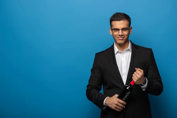 Schöner Geschäftsmann mit einer Flasche Wein in der Hand und lächelnd in die Kamera auf blauem Hintergrund — Stockfoto