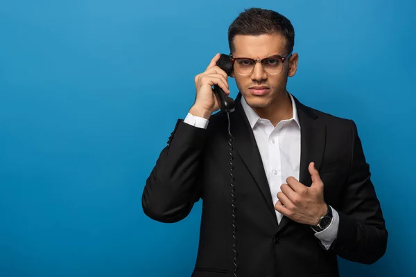 Hombre de negocios confundido mirando a la cámara mientras habla por teléfono sobre fondo azul - foto de stock