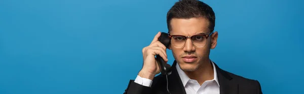 Panoramaaufnahme eines verwirrten Geschäftsmannes, der beim Telefonieren isoliert auf blau in die Kamera schaut — Stockfoto