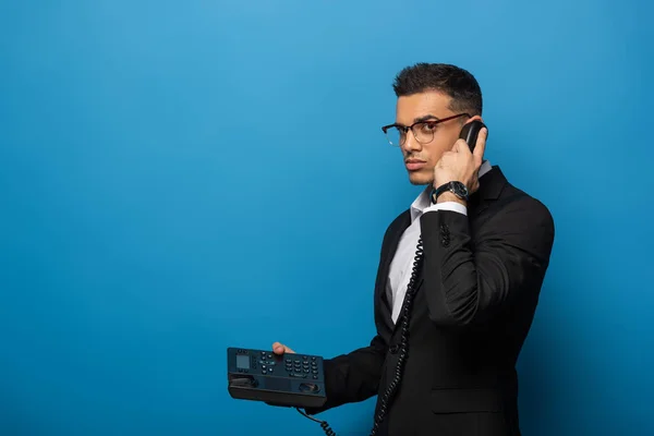 Vista lateral del hombre de negocios hablando por teléfono y mirando a la cámara sobre fondo azul - foto de stock