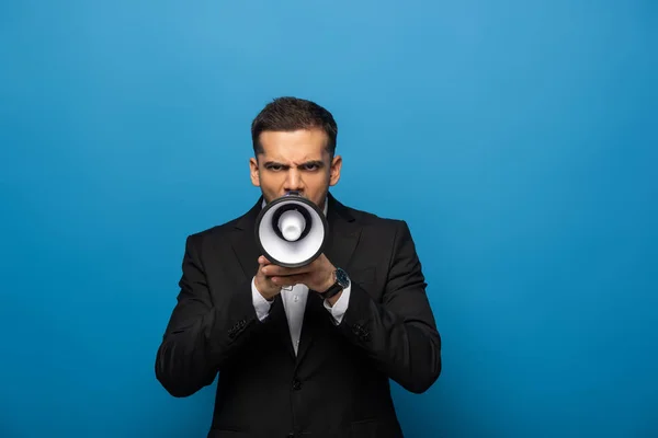 Empresario gritando en altavoz y mirando a la cámara sobre fondo azul - foto de stock