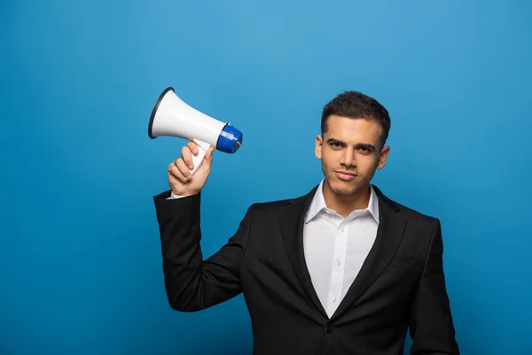 Handsome businessman looking at camera while holding loudspeaker on blue background — Stock Photo
