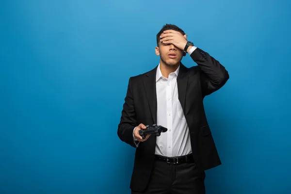 KYIV, UKRAINE - NOVEMBER 12, 2019: Stressed businessman with hand on forehead holding joystick on blue background — Stock Photo