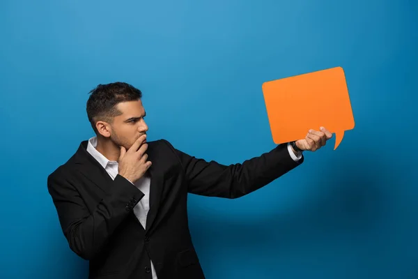 Vista lateral del hombre de negocios reflexivo con la mano por la barbilla que sostiene la burbuja del discurso en el fondo azul - foto de stock