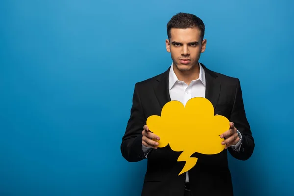 Confident businessman holding speech bubble and looking at camera on blue background — Stock Photo