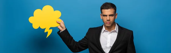 Panoramic shot of businessman with speech bubble looking at camera on blue background — Stock Photo