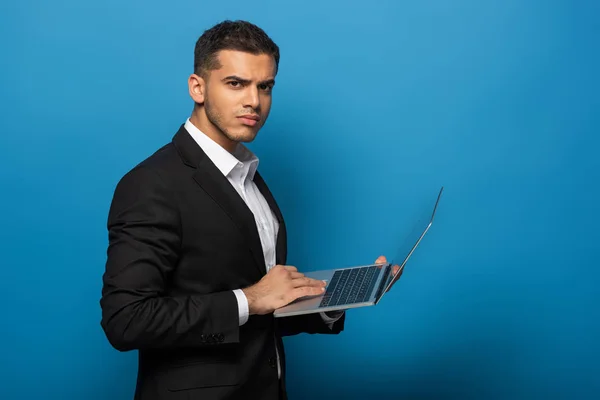 Vista lateral del hombre de negocios utilizando el ordenador portátil y mirando a la cámara en el fondo azul - foto de stock