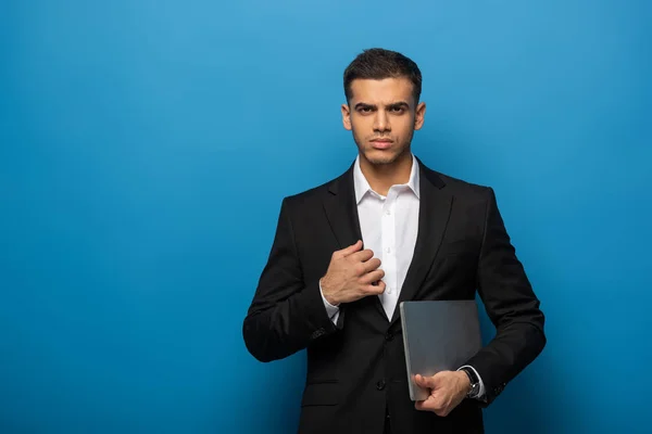 Красивый бизнесмен с ноутбуком смотрит в камеру на синем фоне — стоковое фото
