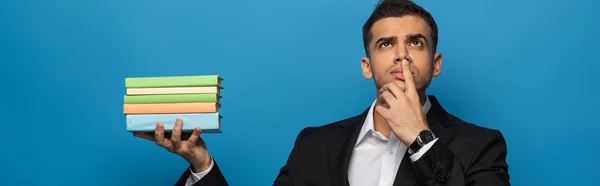 Panoramic shot of pensive businessman holding books and looking away isolated on blue — Stock Photo