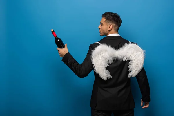 Vista trasera del hombre de negocios con alas blancas sosteniendo botella de vino sobre fondo azul - foto de stock