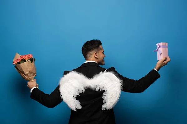 Vista trasera del hombre de negocios con alas blancas sosteniendo regalo y ramo sobre fondo azul - foto de stock