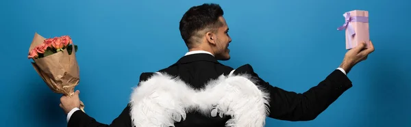 Back view of businessman with white wings holding bouquet and gift on blue background, panoramic shot — Stock Photo