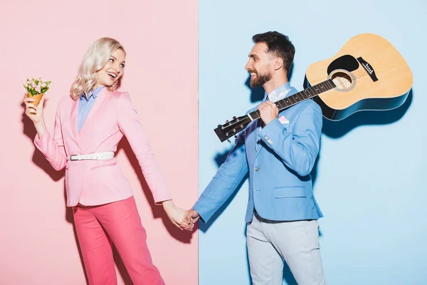 Lächelnde Frau mit Blumenstrauß und gutaussehender Mann mit Akustikgitarre an der Hand auf rosa und blauem Hintergrund — Stockfoto