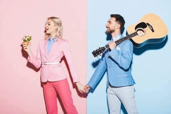 Schockierte Frau mit Blumenstrauß und gutaussehender Mann mit Akustikgitarre, der Hände auf rosa und blauem Hintergrund hält — Stockfoto