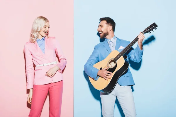 Усміхнена жінка і красивий чоловік грає на акустичній гітарі на рожевому і синьому фоні — стокове фото