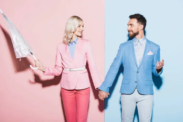 Lächelnde Frau mit Regenschirm und gutaussehender Mann, der Hände auf rosa und blauem Hintergrund hält — Stockfoto