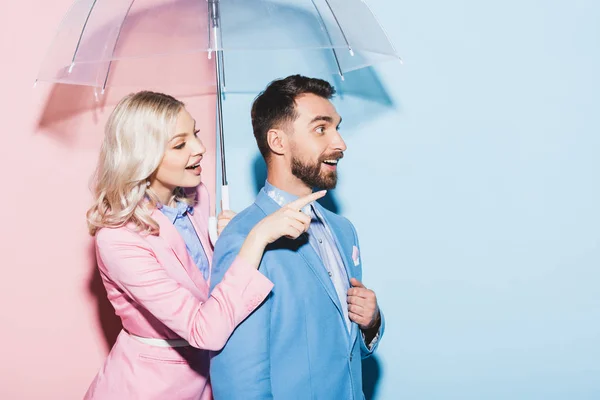 Lächelnde Frau mit Regenschirm, der mit dem Finger zeigt und schockierter Mann auf rosa und blauem Hintergrund — Stockfoto