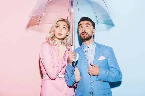 Schockierte Frau und schöner Mann mit Regenschirm auf rosa und blauem Hintergrund — Stockfoto