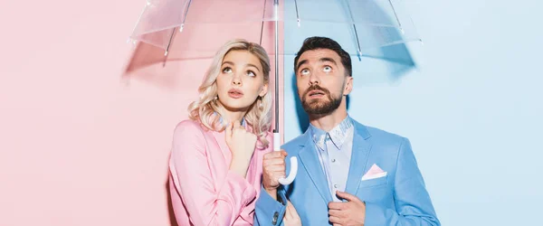 Panoramic shot of shocked woman and handsome man with umbrella looking up on pink and blue background — Stock Photo