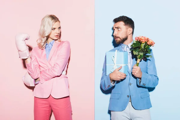 Serious woman with boxing gloves and scared man with gift and bouquet on pink and blue background — Stock Photo