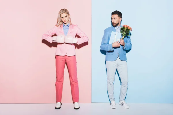 Mujer seria con guantes de boxeo y hombre asustado con regalo y ramo sobre fondo rosa y azul — Stock Photo