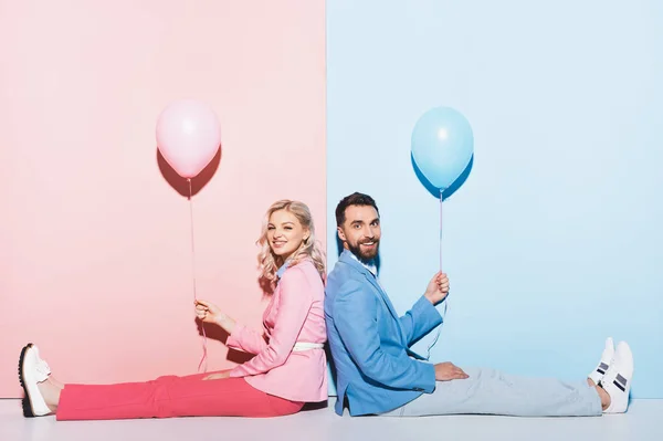 Спина к спине вид улыбающейся женщины и красивого мужчины, держащего воздушные шары на розовом и голубом фоне — стоковое фото