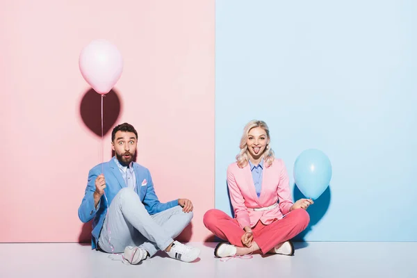 Attraktive Frau streckt die Zunge heraus und schockierter Mann mit Luftballons auf rosa und blauem Hintergrund — Stockfoto