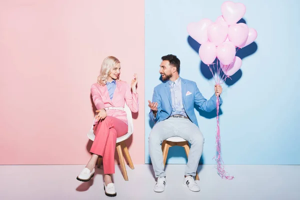 Mujer sonriente mirando al hombre guapo con globos sobre fondo rosa y azul - foto de stock