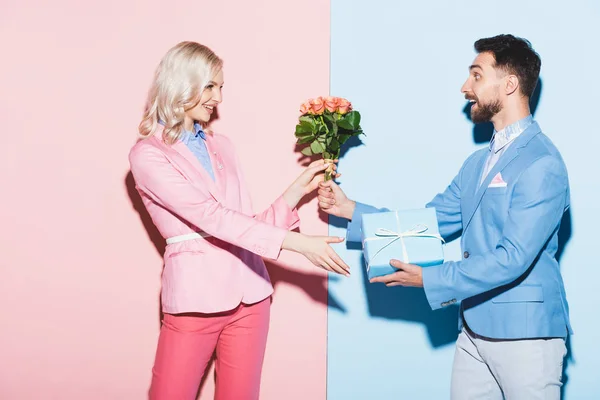 Schöner Mann schenkt lächelnden Frau Strauß und Geschenk auf rosa und blauem Hintergrund — Stockfoto