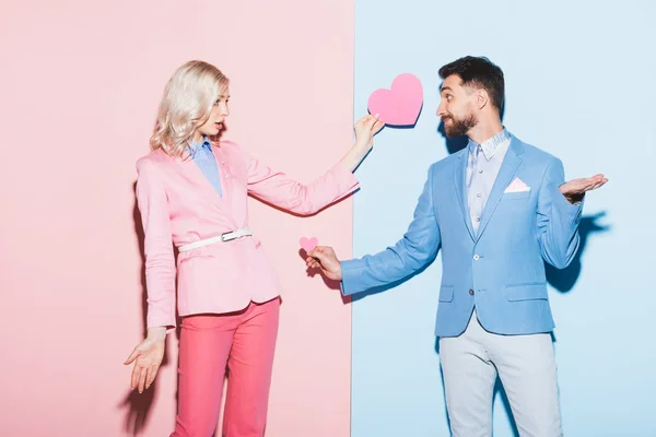 Shocked woman and handsome man holding heart-shaped cards on pink and blue background — Stock Photo