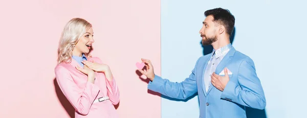 Colpo panoramico di bell'uomo che dà carta a forma di cuore alla donna scioccata su sfondo rosa e blu — Foto stock