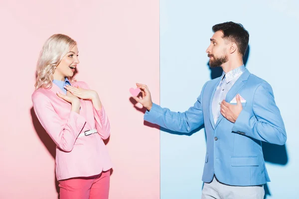 Schöner Mann gibt einer schockierten Frau eine herzförmige Karte auf rosa und blauem Hintergrund — Stockfoto