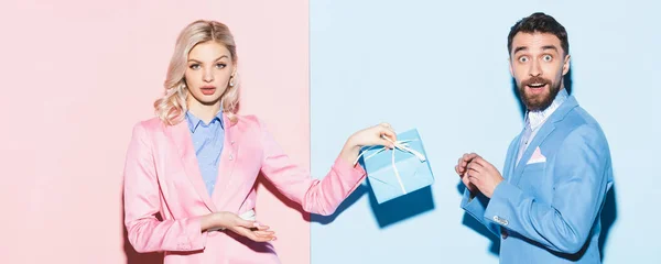 Panoramic shot of shocked woman holding gift and handsome man on pink and blue background — Stock Photo