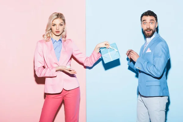 Schockierte Frau mit Geschenk und gutaussehendem Mann auf rosa und blauem Hintergrund — Stockfoto