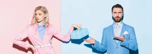 Panoramic shot of sad woman holding gift and shocked man pointing with hands on pink and blue background — Stock Photo