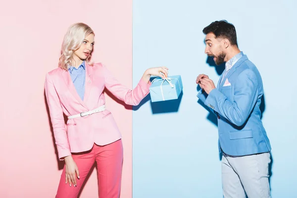 Attractive woman giving present to shocked man on pink and blue background — Stock Photo
