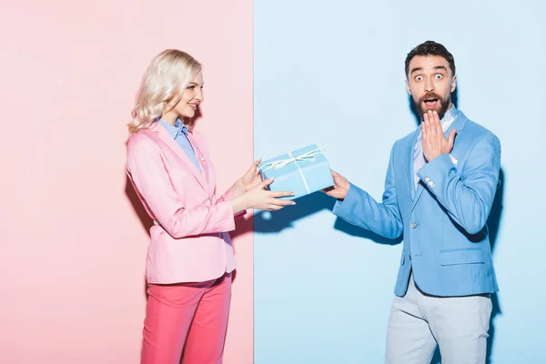 Femme souriante donnant cadeau à l'homme choqué sur fond rose et bleu — Photo de stock