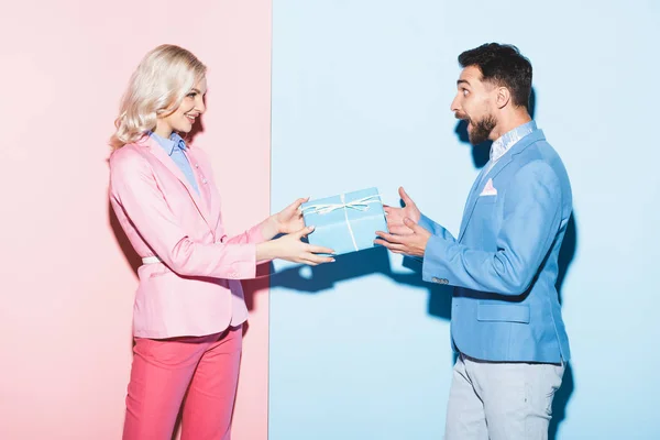 Vue latérale de la femme souriante donnant cadeau à l'homme choqué sur fond rose et bleu — Photo de stock