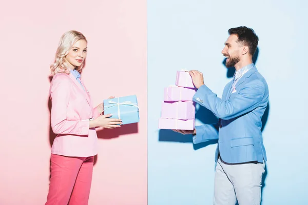 Attractive woman and smiling man holding gifts on pink and blue background — Stock Photo