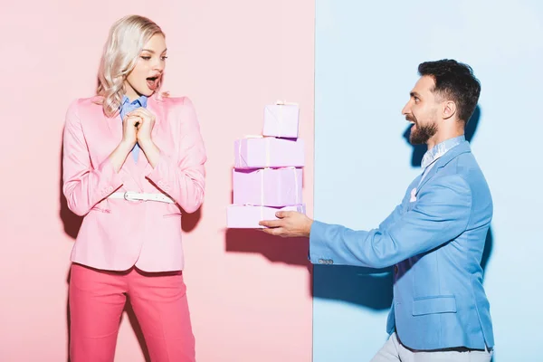 Handsome man giving gifts to shocked woman on pink and blue background — Stock Photo